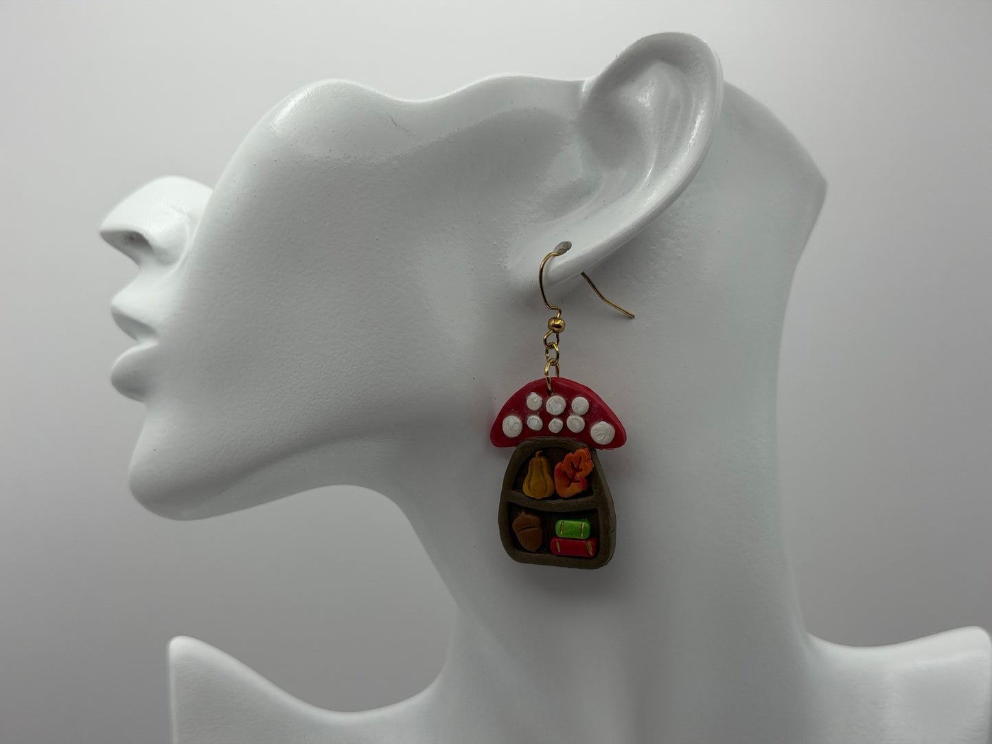 A pair of whimsical fairy house earrings displayed on a white mannequin head. The earrings feature a red and white mushroom cap with white polka dots and a small brown fairy house filled with tiny colorful accessories.