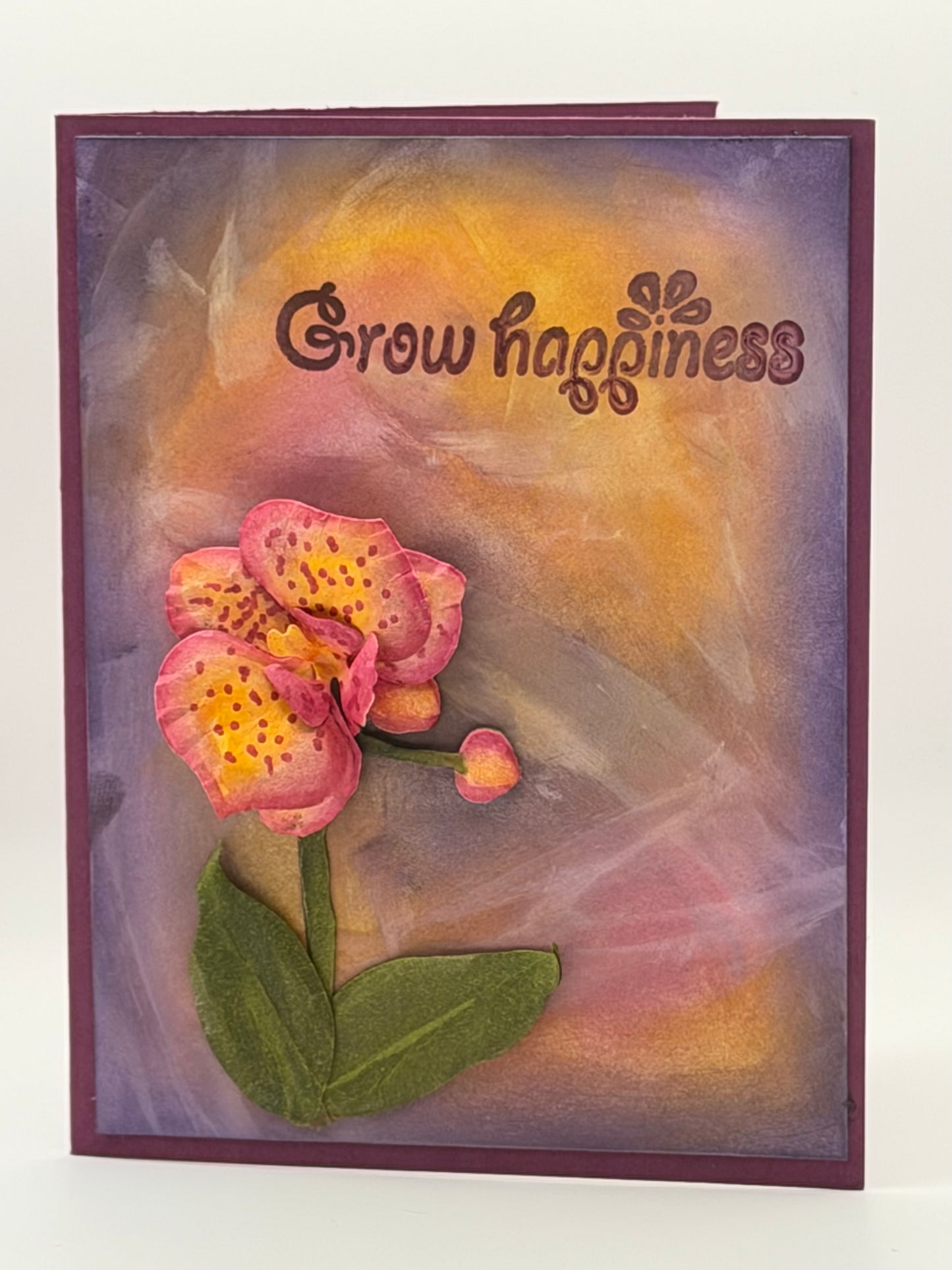 A handmade greeting card featuring a textured pink and yellow orchid flower with green leaves against a purple and orange abstract background, with the words 'Grow happiness' stamped on the top.
