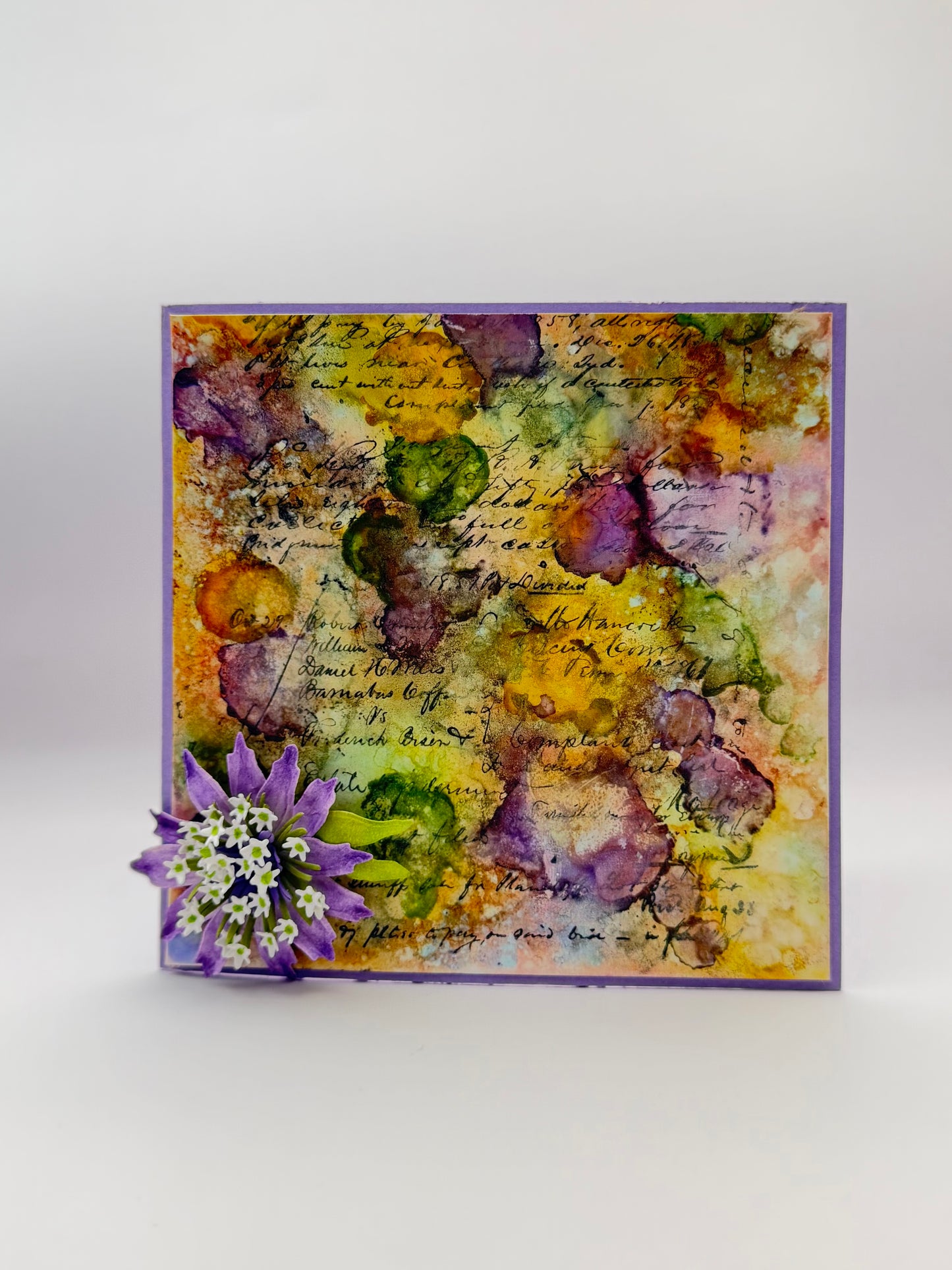 A square handmade greeting card with a purple border, featuring an abstract watercolor design in yellow, green, and purple with cursive script, decorated with a small purple flower on the bottom left, set against a light gray background.