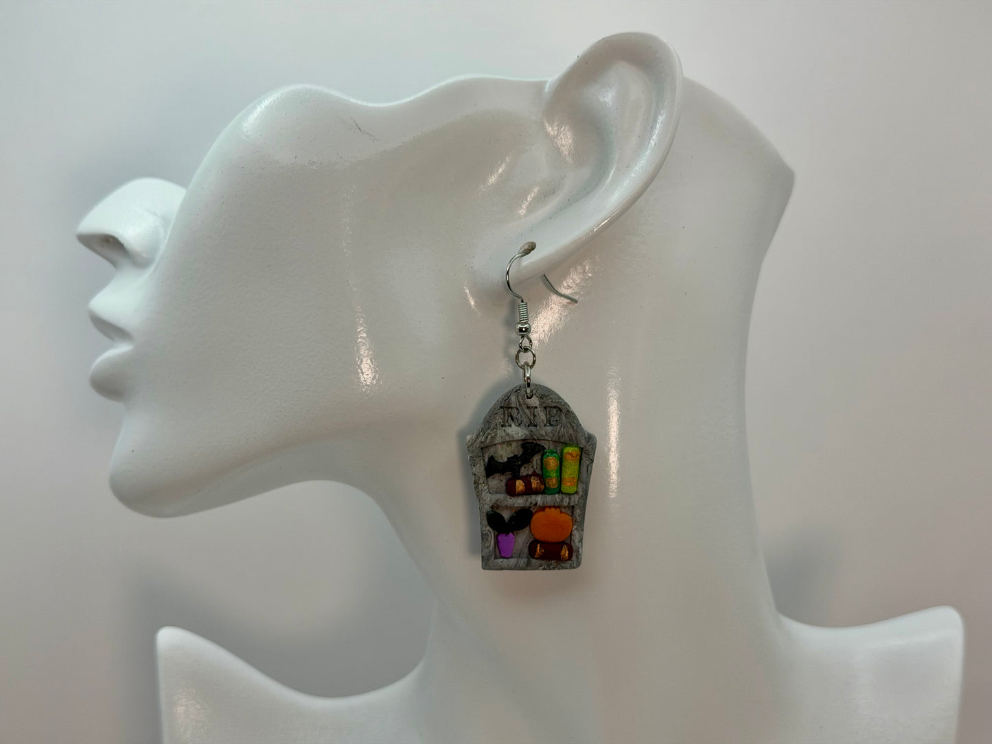 A single dangling earring displayed on a white mannequin ear. The earring features a gray tombstone bookshelf earring with small colorful Halloween-themed details, including a black bat, green books, plant, and an orange pumpkin, hanging from a silver fishhook. Charmedandcrowned.com