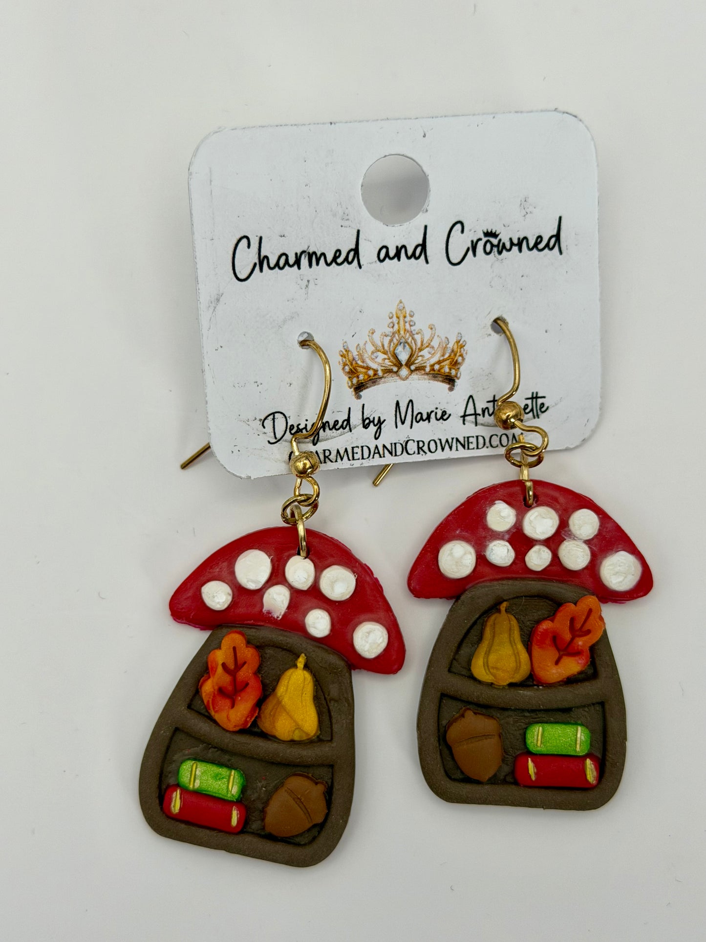 A pair of mushroom-shaped dangling earrings displayed on a white branded card. The earrings have red caps with white spots, brown bodies, and small autumn-themed details including orange leaves, yellow pears, green books, and brown acorns. CharmedandCrowned.com
