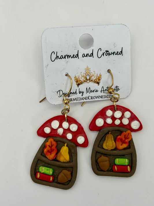 A pair of mushroom-shaped dangling earrings displayed on a white branded card. The earrings have red caps with white spots, brown bodies, and small autumn-themed details including orange leaves, yellow pears, green books, and brown acorns. CharmedandCrowned.com