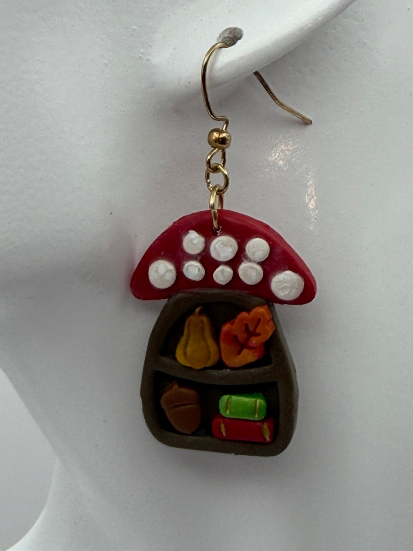 A close-up of a handmade polymer clay earring featuring a red mushroom cap with white spots and a dark brown compartmentalized tray with small orange, yellow, green, and brown details, attached to a gold-tone fishhook ear wire.