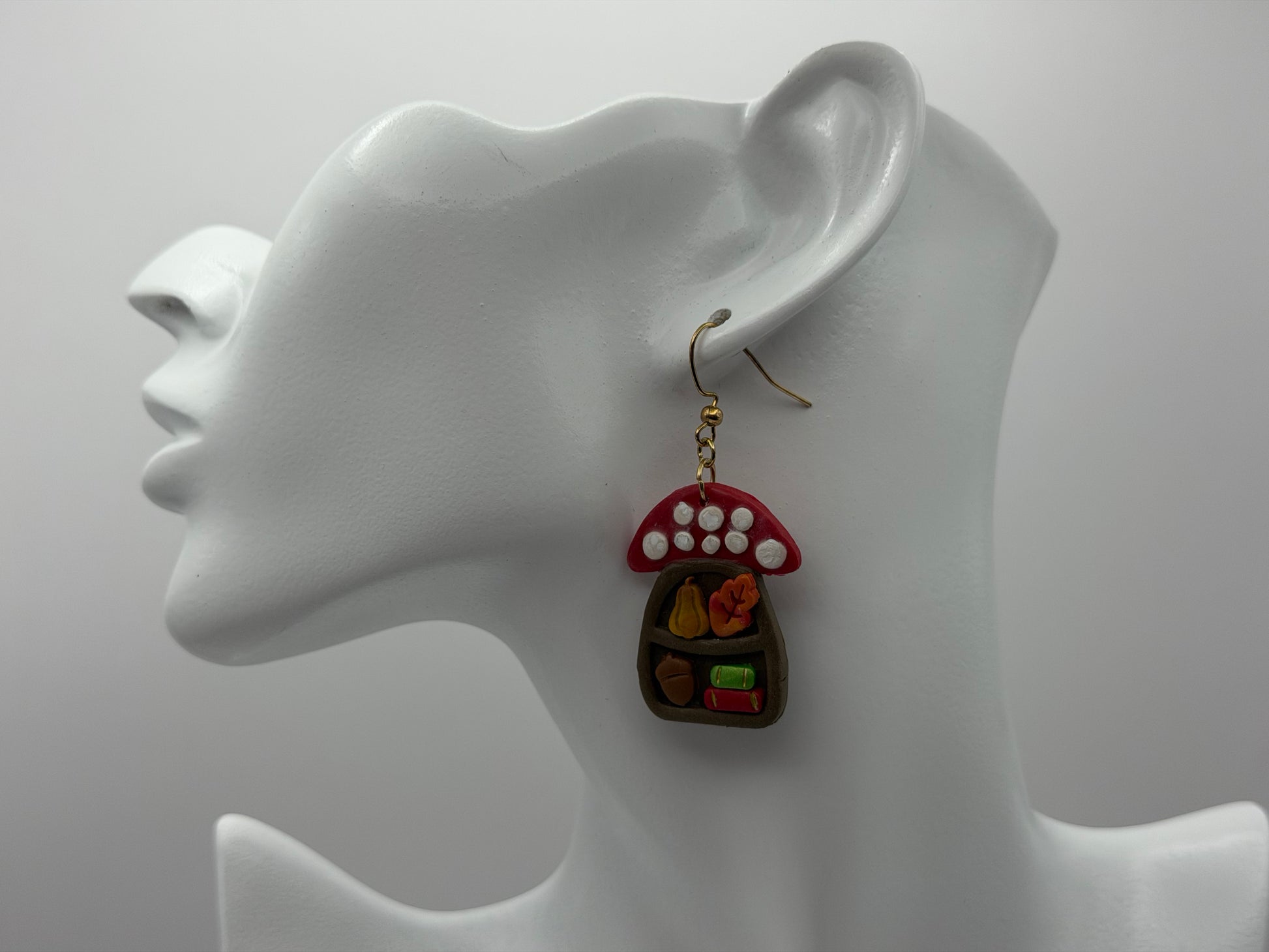 A pair of whimsical fairy house earrings displayed on a white mannequin head. The earrings feature a red and white mushroom cap with white polka dots and a small brown fairy house filled with tiny colorful accessories.