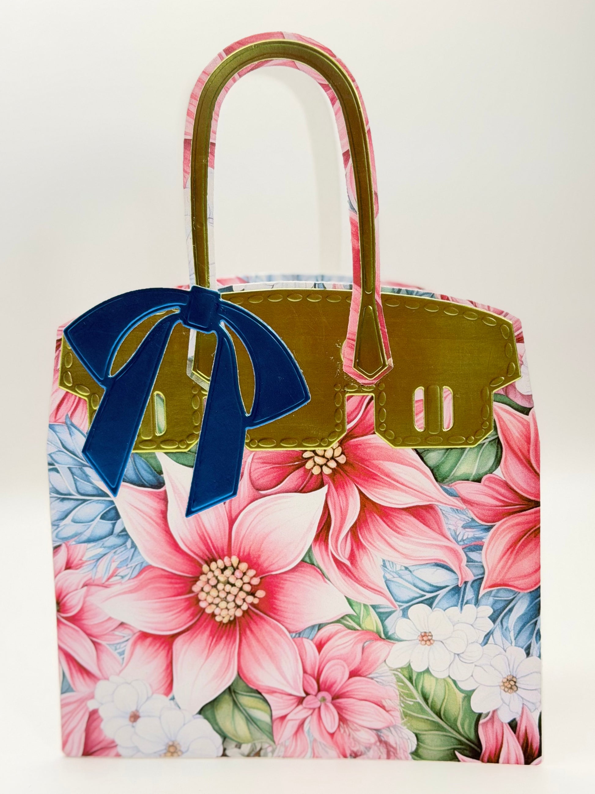 A structured handbag with a pink and blue floral print, olive green trim, and a large navy blue bow on the front, photographed against a plain light background. Charmedandcrowned.com