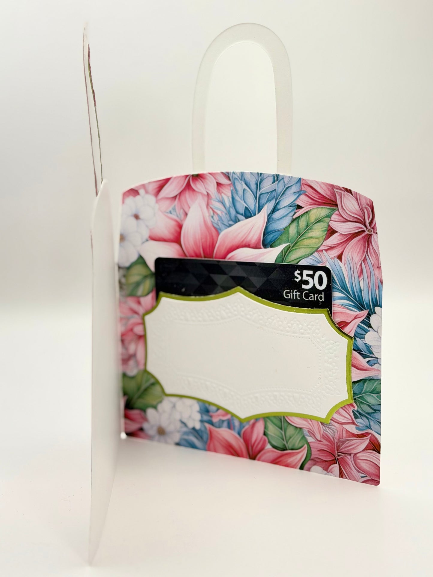 A gift card in a decorative paper bag packaging with a pink, blue, and green floral pattern, featuring a white plastic handle. Charmedandcrowned.com