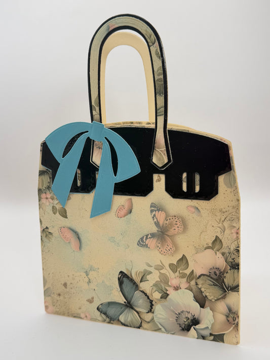 A decorative paper bag styled to look like a handbag, featuring a cream and beige background with a butterfly and floral print, black trim, and a light blue bow on the front.