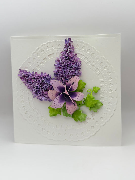 A square white card featuring a raised circular lace-like pattern, decorated with three artificial purple and lavender flowers and green leaves, photographed against a neutral light gray background.