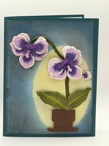 A handmade greeting card featuring a 3D layered design of purple and lavender orchids in a brown pot against a blue textured background with a yellow oval glow.