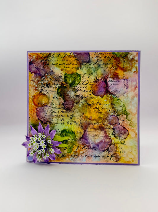 A square handmade greeting card with a purple border, featuring an abstract watercolor design in yellow, green, and purple with cursive script, decorated with a small purple flower on the bottom left, set against a light gray background.