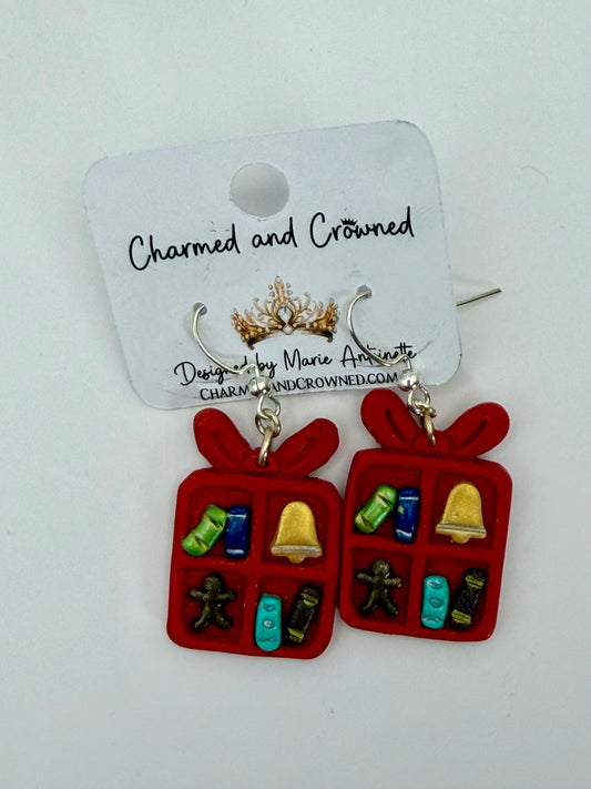 A pair of red gift box dangle earrings with small holiday-themed charms inside, hanging from silver hooks on a white display card that reads 'Charmed and Crowned'.