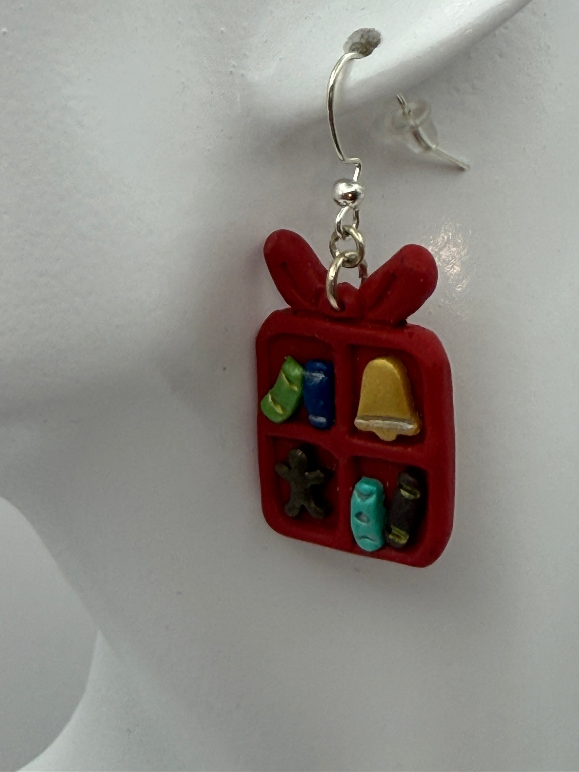 A pair of red gift box dangle earrings with small holiday-themed charms inside, hanging from silver hooks on a white display card that reads 'Charmed and Crowned'.