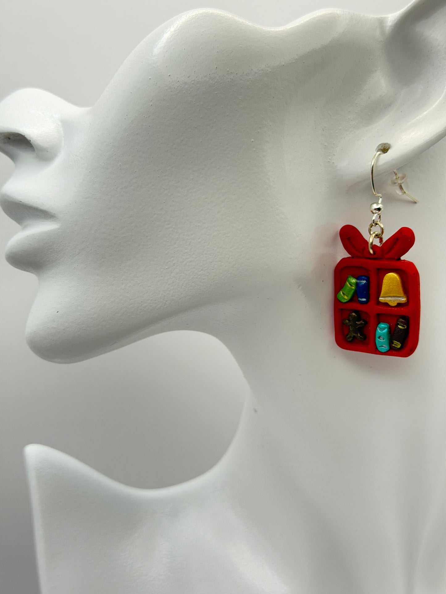 A pair of red gift box dangle earrings with small holiday-themed charms inside, hanging from silver hooks on a white display card that reads 'Charmed and Crowned'.