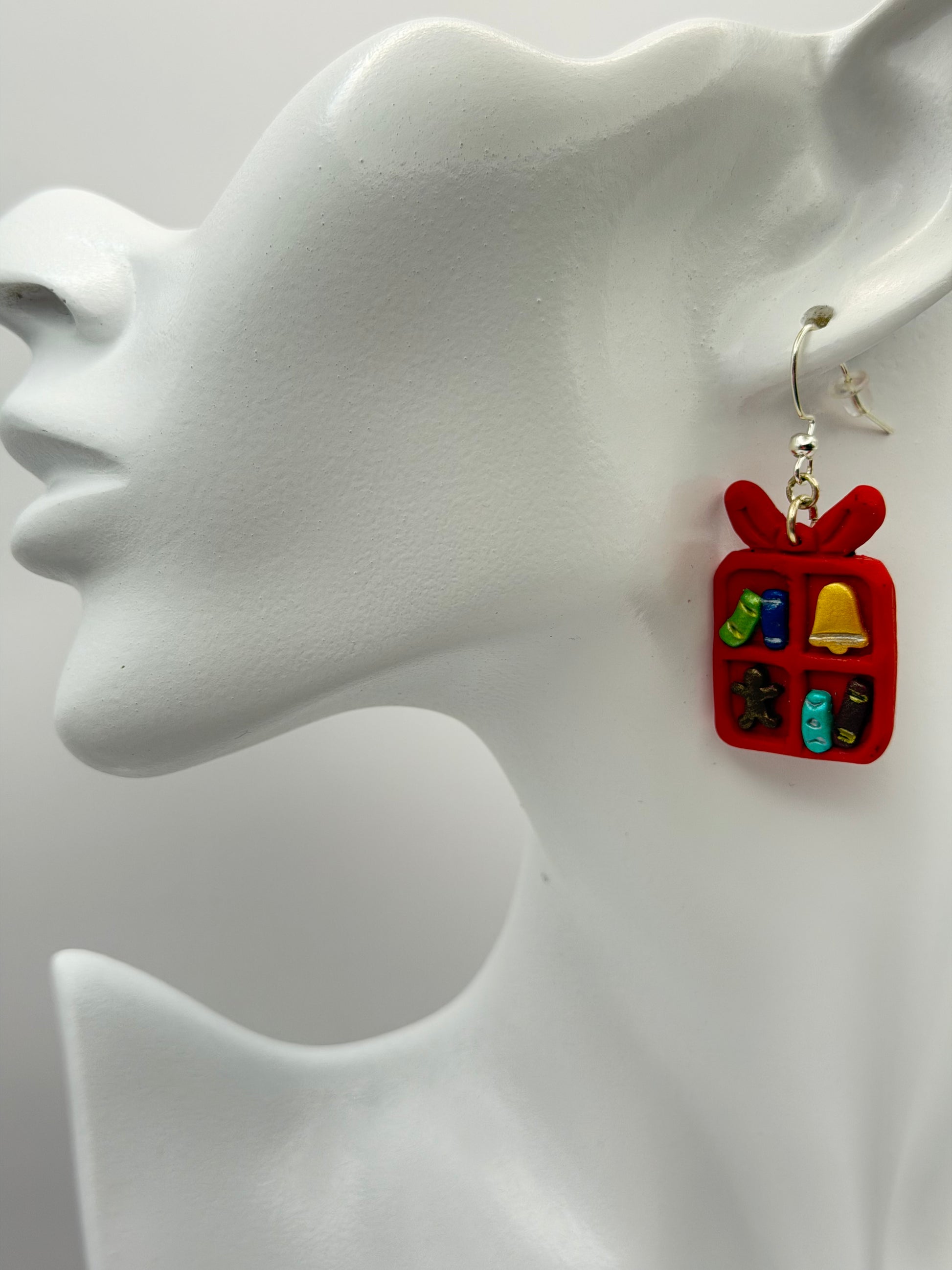 A pair of red gift box dangle earrings with small holiday-themed charms inside, hanging from silver hooks on a white display card that reads 'Charmed and Crowned'.