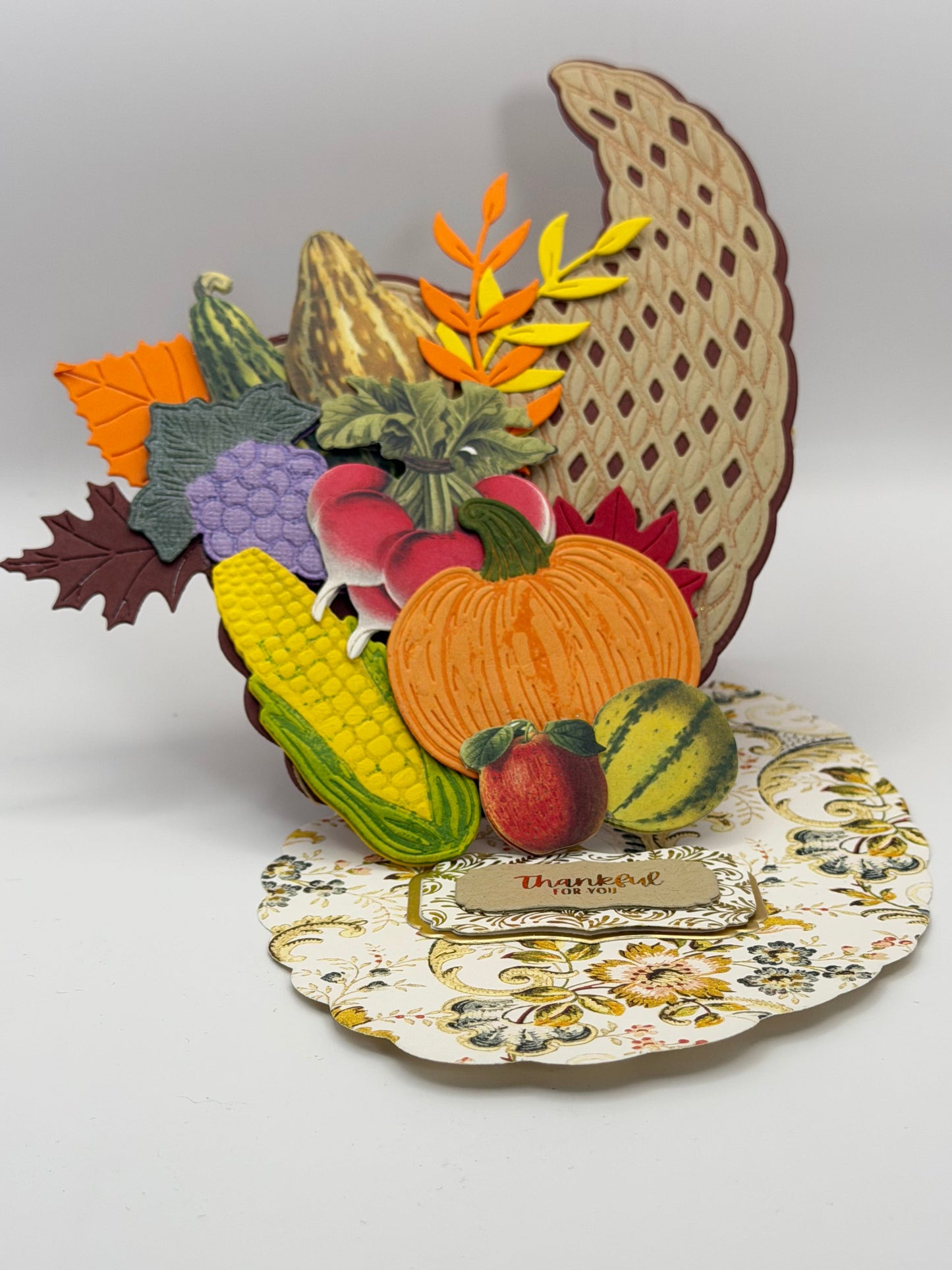 A decorative autumn-themed wall hanging featuring layered paper cutouts of a cornucopia with pumpkins, corn, grapes, leaves, and a floral plate on a plain light background.