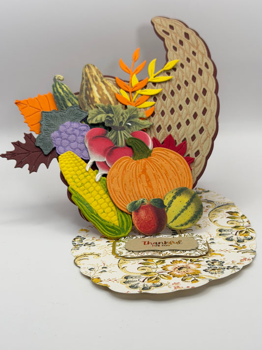 A decorative autumn-themed wall hanging featuring layered paper cutouts of a cornucopia with pumpkins, corn, grapes, leaves, and a floral plate on a plain light background.