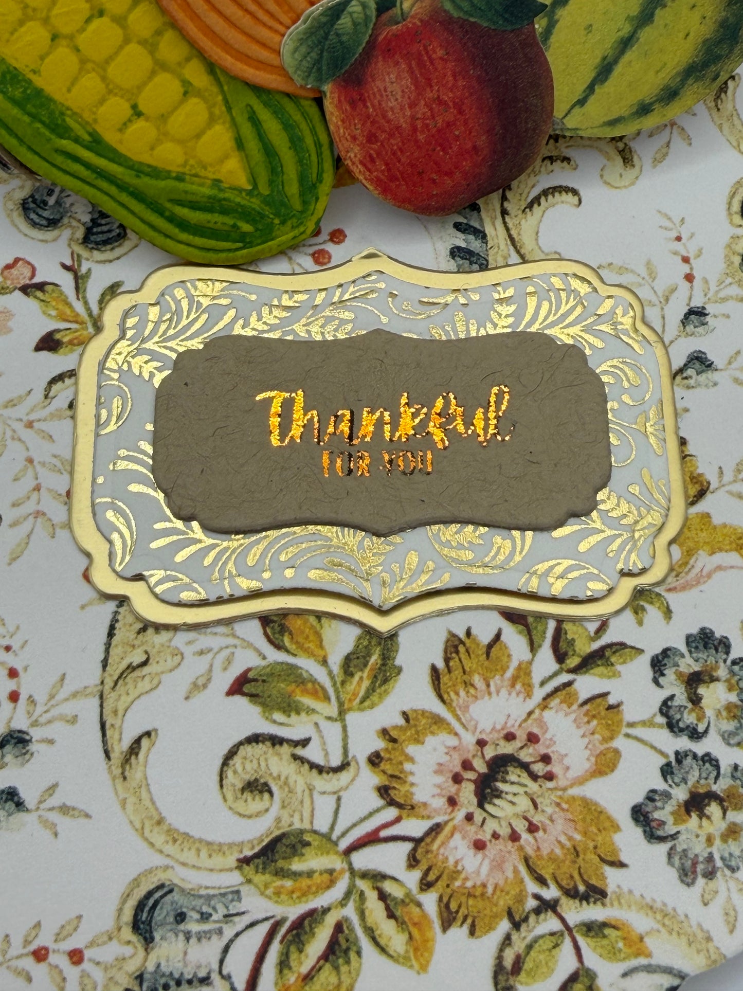 A decorative scalloped card or label with a gray center and gold foil border, featuring the handwritten text 'Thankful for who you are,' placed on a floral surface with autumn-themed props partially visible at the left edge.