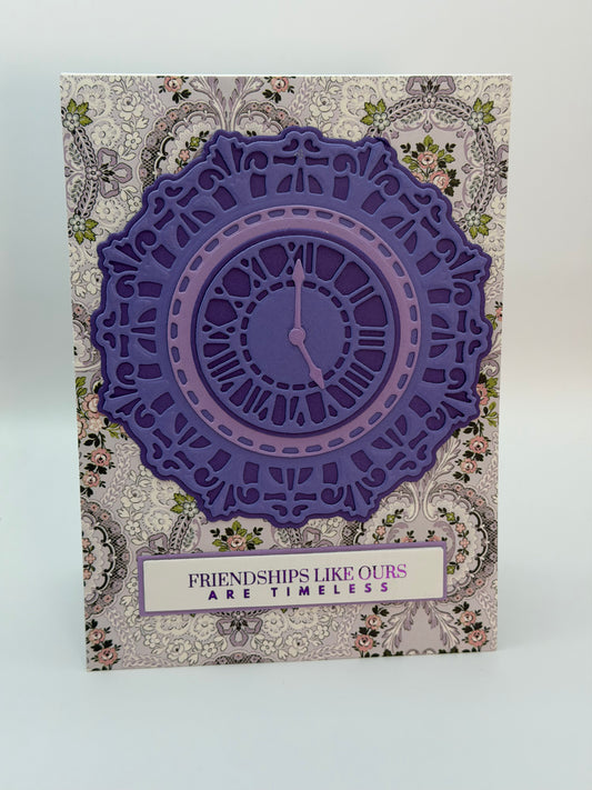 A rectangular decorative card or art print featuring a large layered purple clock design over a beige and gray floral paisley background, with a vertical white label reading 'FRIENDSHIPS LIKE OURS ARE TIMELESS'.