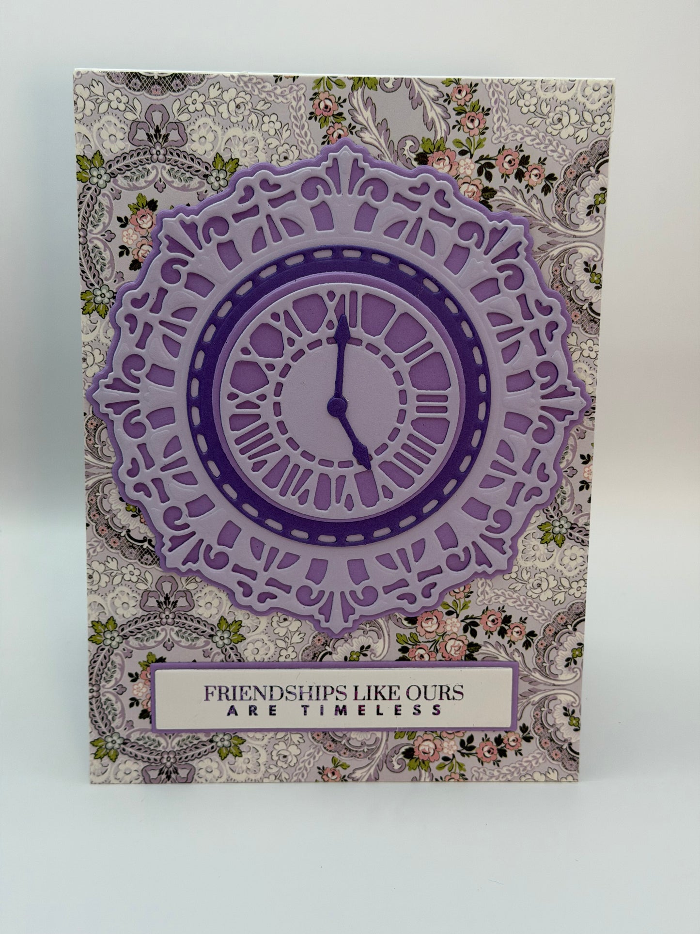 A square greeting card featuring a layered purple clock design over a vintage floral and paisley patterned background, with the text 'FRIENDSHIPS LIKE OURS ARE TIMELESS' at the bottom.