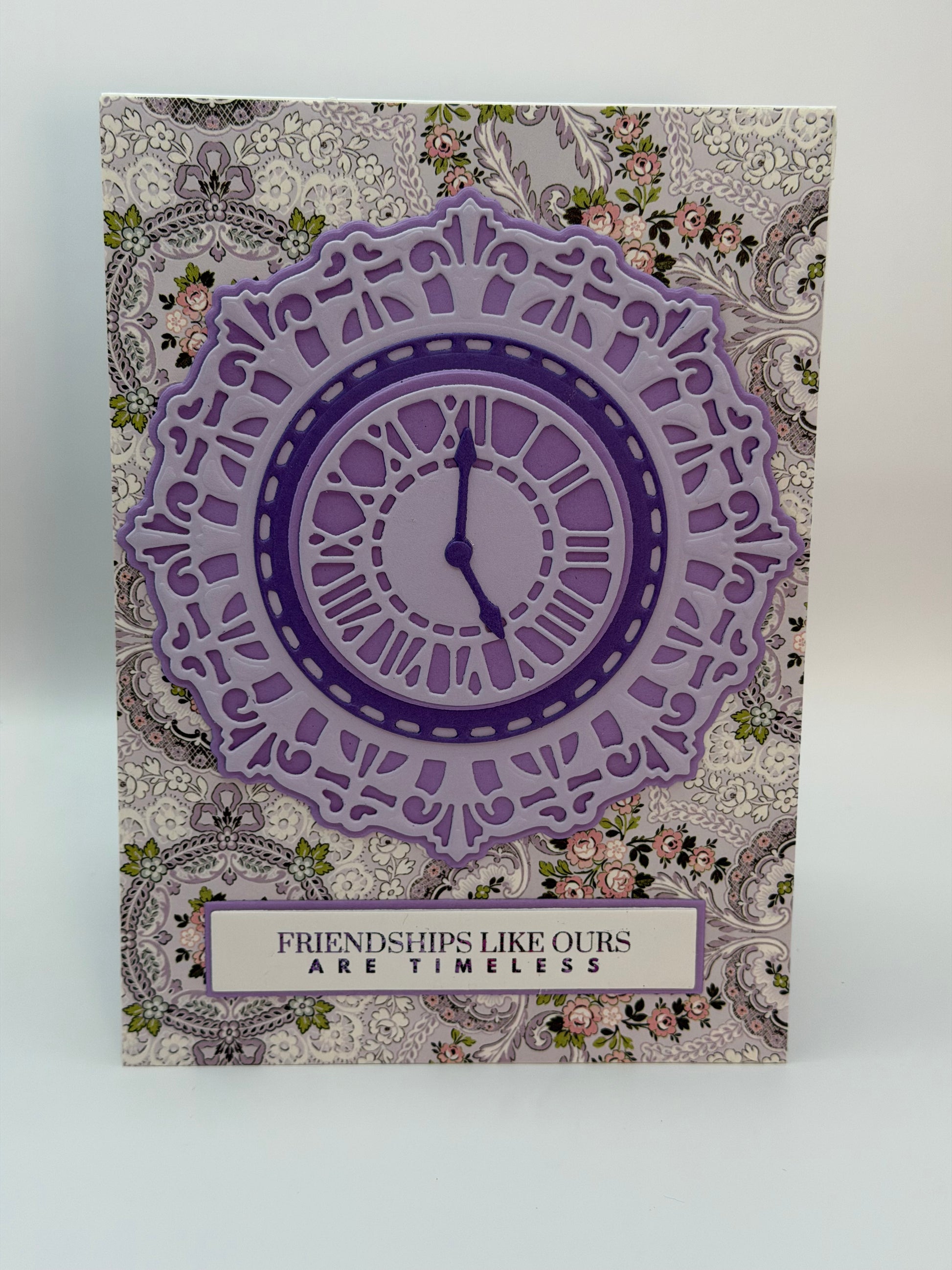 A square greeting card featuring a layered purple clock design over a vintage floral and paisley patterned background, with the text 'FRIENDSHIPS LIKE OURS ARE TIMELESS' at the bottom.
