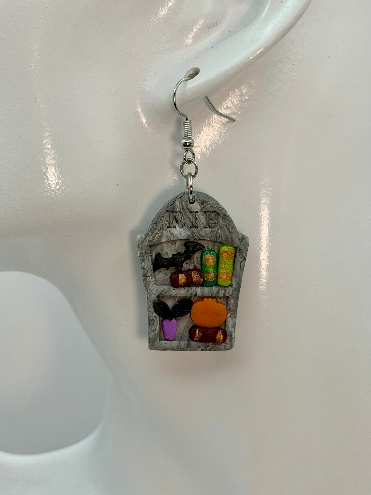 A single dangling earring displayed on a white mannequin ear. The earring features a gray tombstone bookshelf earring with small colorful Halloween-themed details, including a black bat, green books, plant, and an orange pumpkin, hanging from a silver fishhook. Charmedandcrowned.com