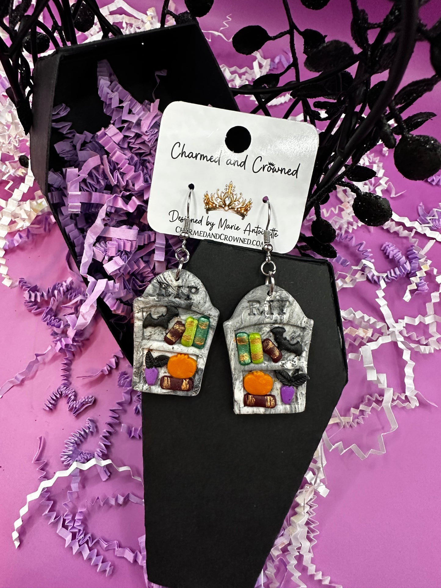 A single dangling earring displayed on a white mannequin ear. The earring features a gray tombstone bookshelf earring with small colorful Halloween-themed details, including a black bat, green books, plant, and an orange pumpkin, hanging from a silver fishhook. Charmedandcrowned.com